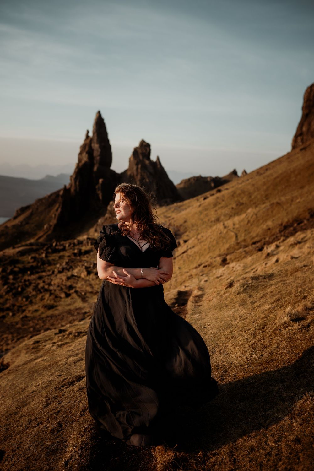 A flowing black dress catches the wind against dramatic rocky spires of the Old Man of Storr in Scotland at sunset.