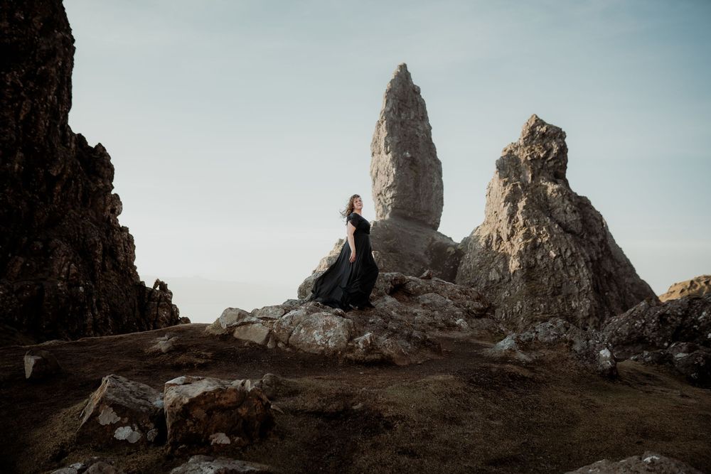 A silhouette sits on rocky cliffs near the towering Old Man of Storr formation on Isle of Skye in Scotland at sunset.