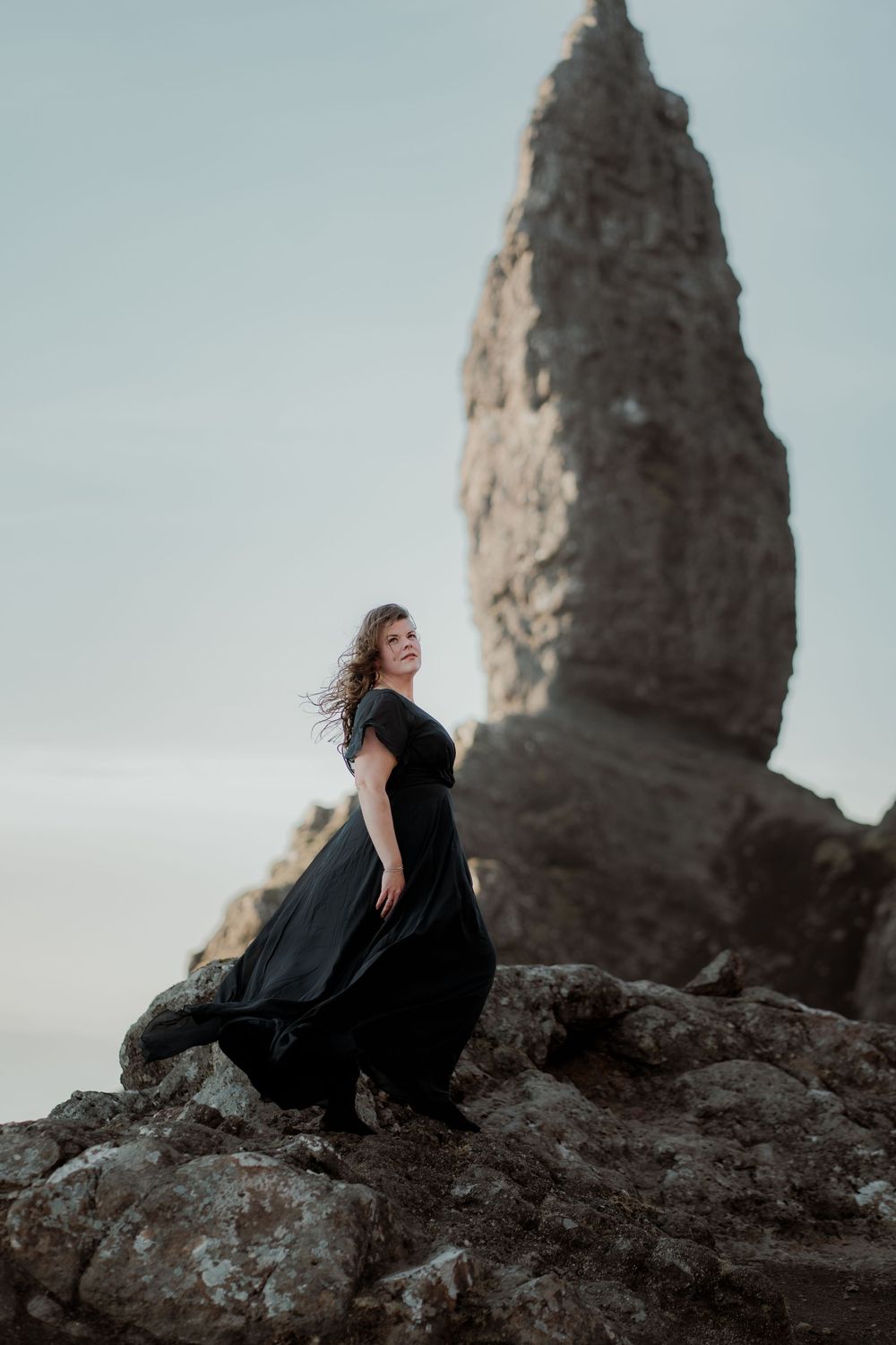 A flowing black dress billows in the wind as someone stands dramatically on rocky cliffs beside a towering stone spire.
