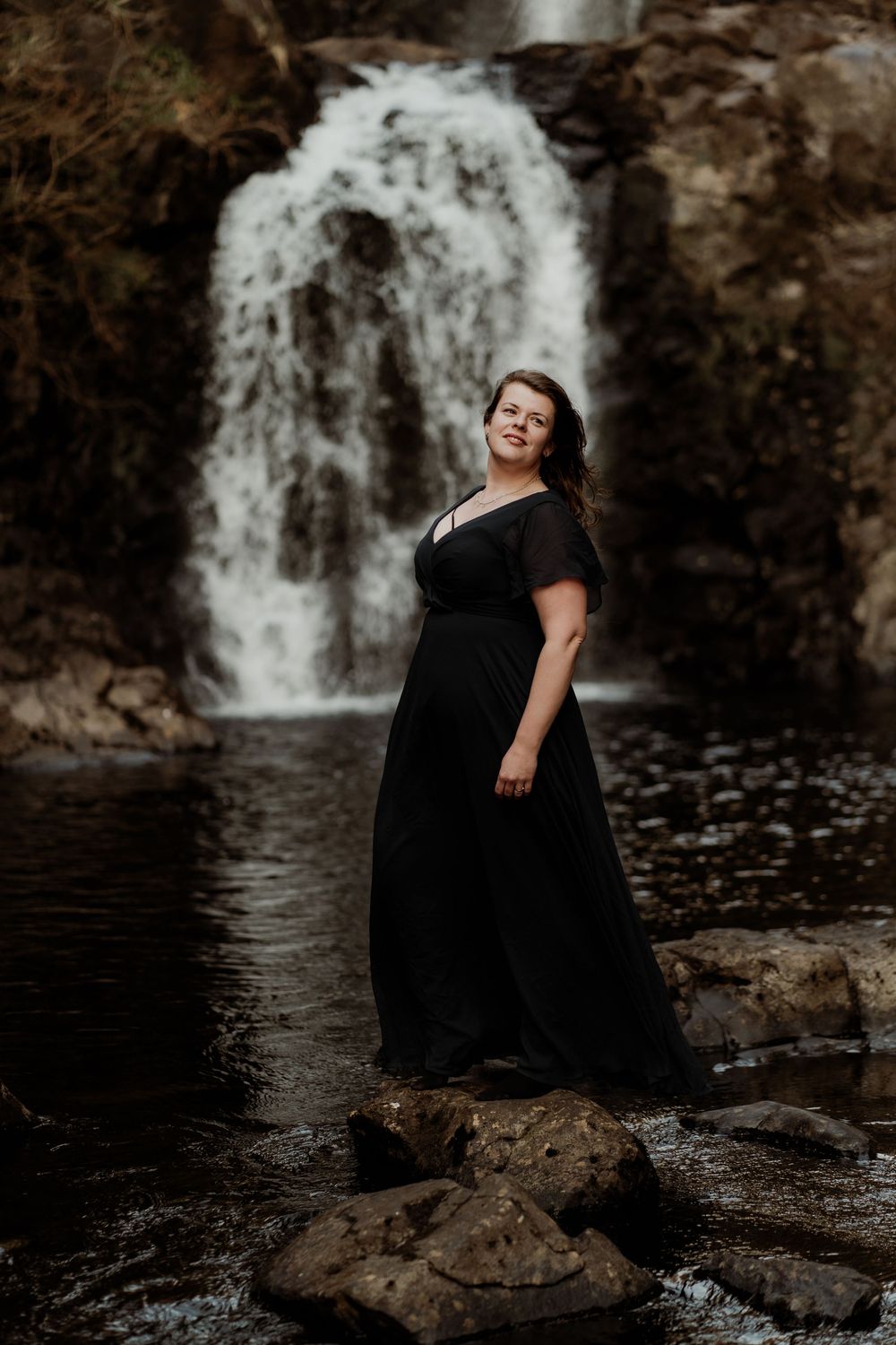 A figure in a long black dress stands on rocks near a cascading waterfall against a moody natural backdrop.