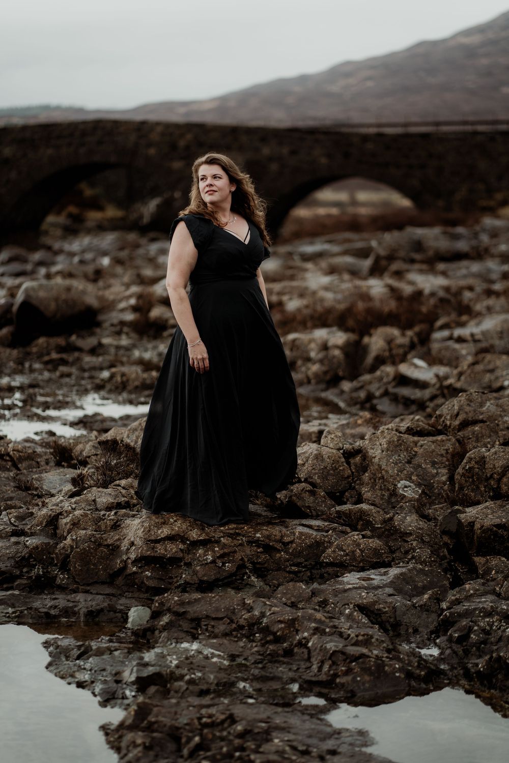 A figure in a flowing black dress stands gracefully on dark volcanic rocks near the ocean at sunset.
