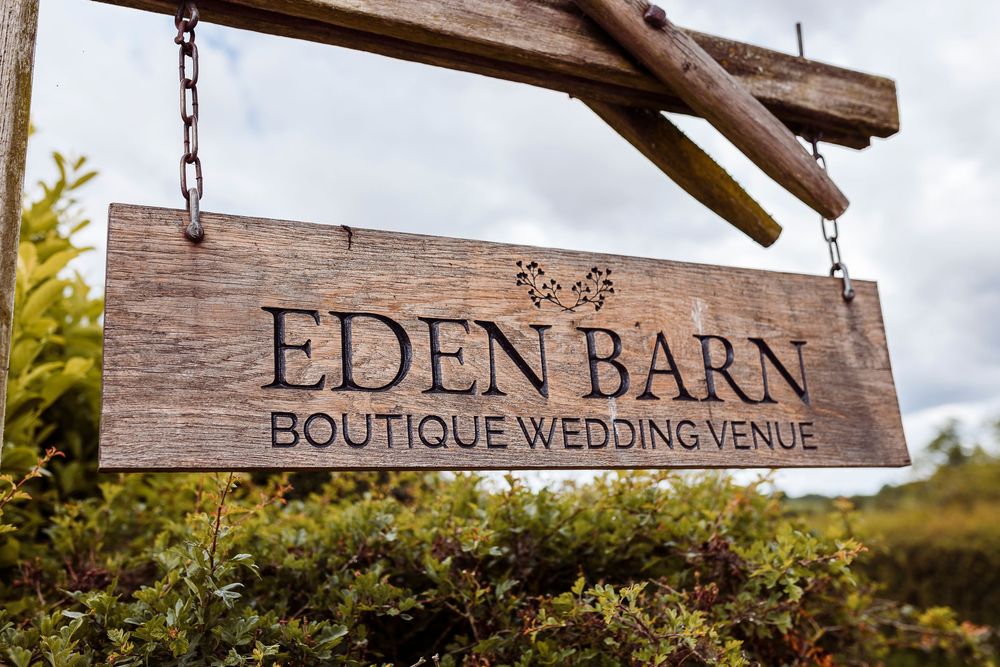 A rustic wooden sign reading 'Eden Barn Boutique Wedding Venue' hangs from chains against a cloudy sky.