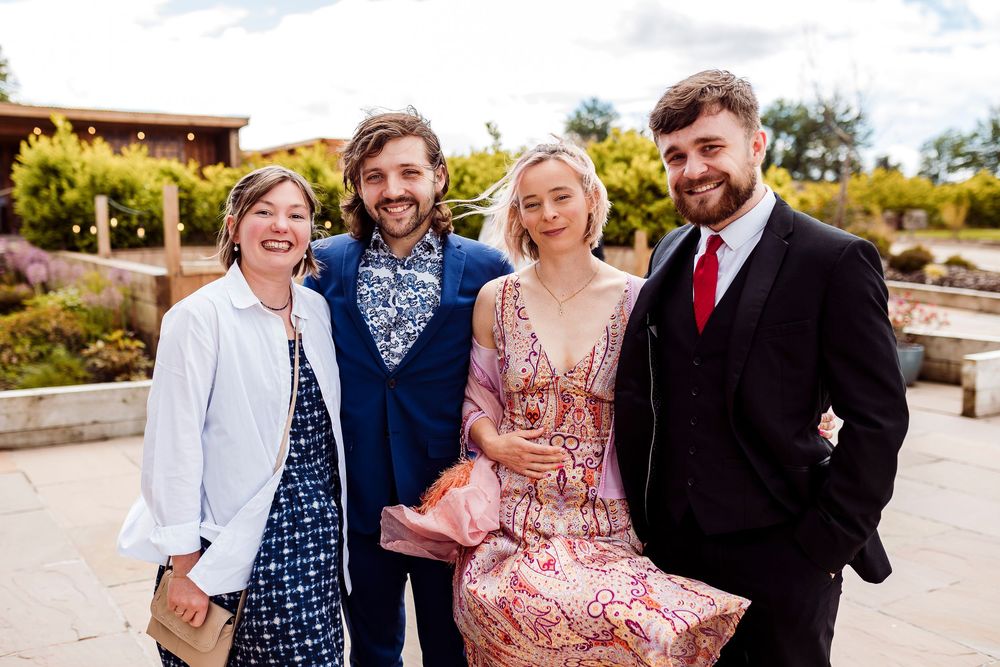 Group of four people posing outdoors in dressy attire with warm smiles and happy expressions on a sunny day.