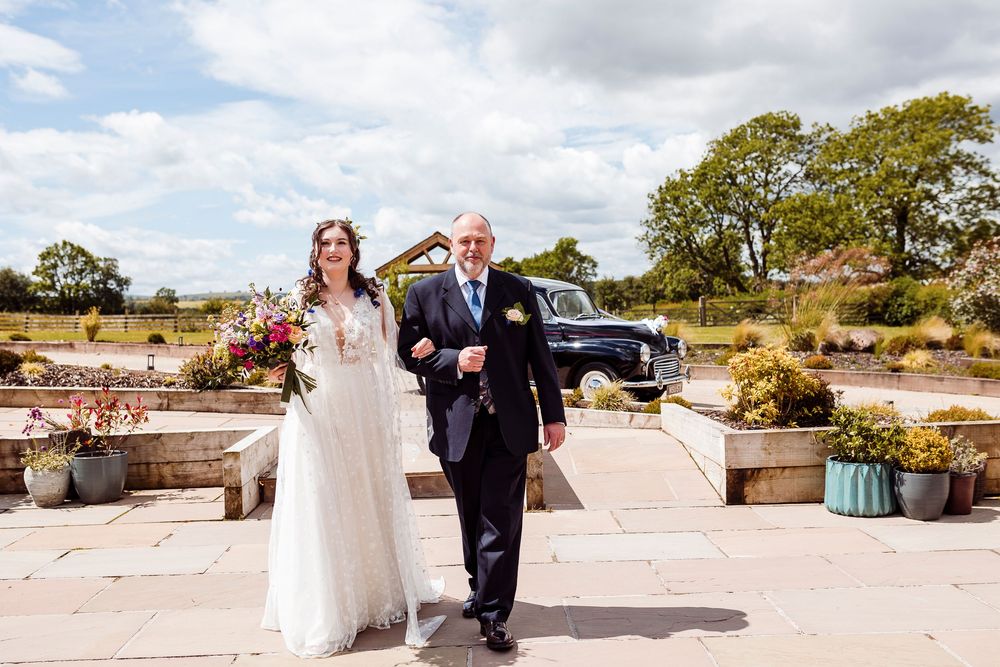 A traditional countryside wedding processional taking place on stone steps surrounded by potted plants on a sunny day.