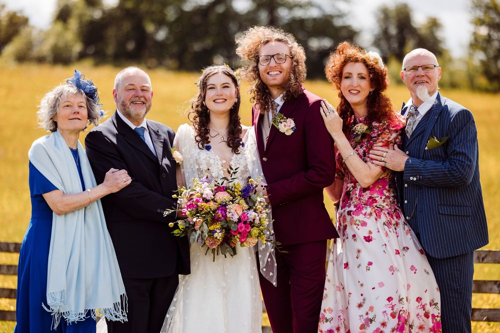 A wedding party in colorful formal attire posing together outdoors in a sunlit field for a group portrait.