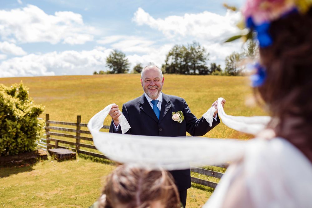 A joyful outdoor wedding moment captured against rolling hills and a wooden fence on a bright summer day.