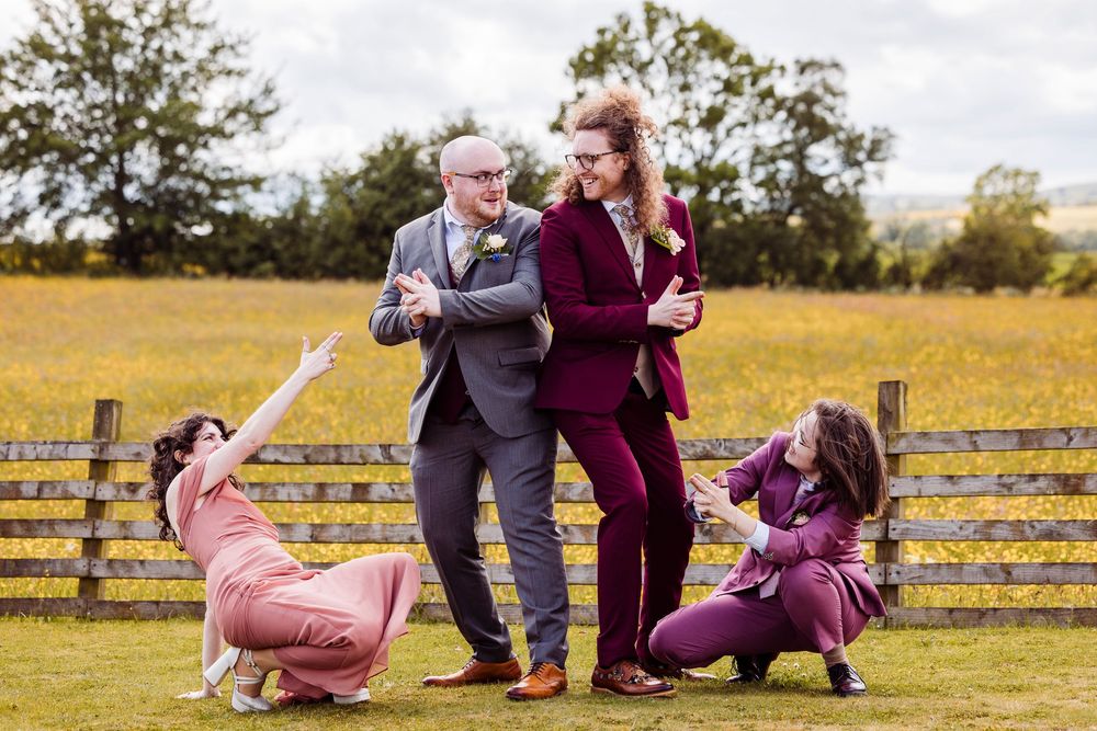Group of people in burgundy and pink outfits strike playful dramatic poses in a field near a rustic wooden fence.