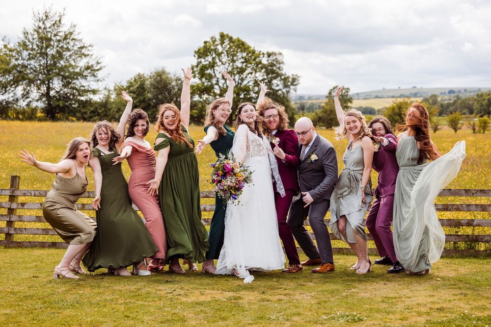 Wedding party poses energetically on grassy hill with rustic wooden fence and trees in background.