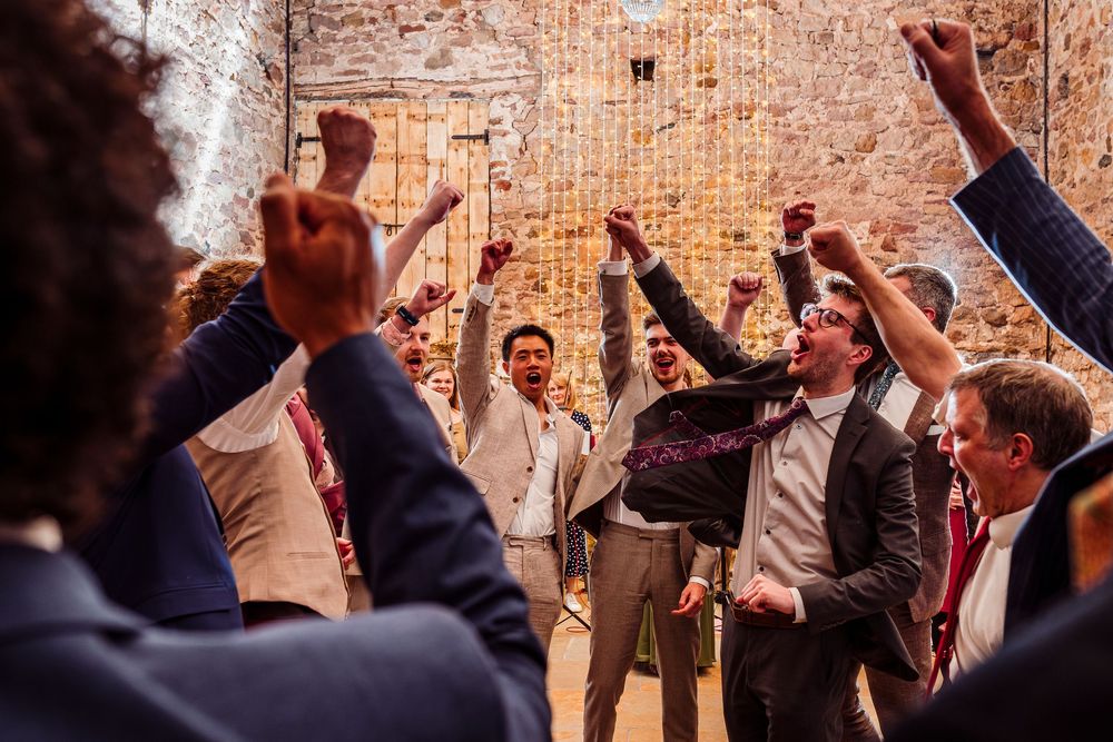 Wedding reception guests celebrate with raised arms on the dance floor in a rustic brick-walled venue during festivities.
