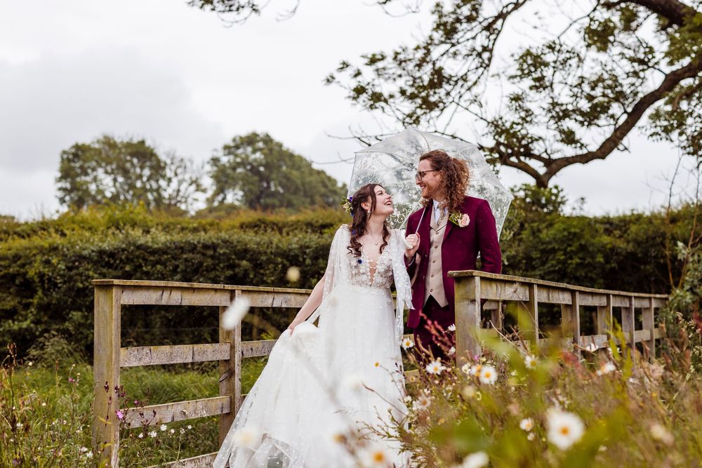 A romantic wedding couple poses by a rustic wooden fence surrounded by wildflowers and greenery.