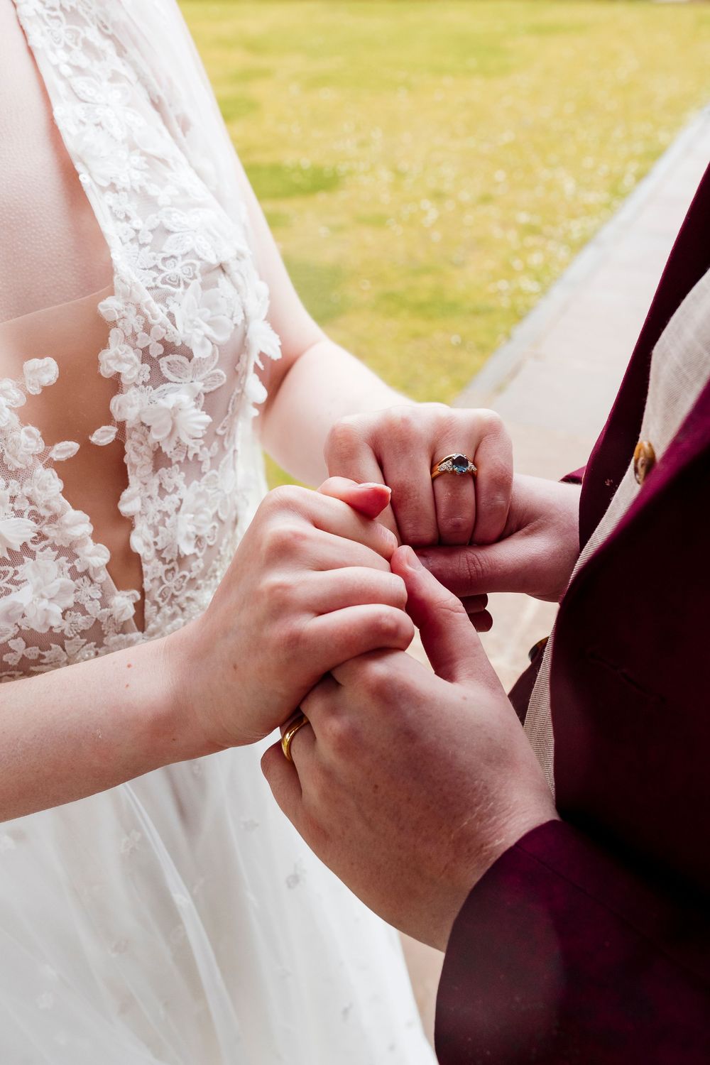 Close-up of hands exchanging wedding rings during ceremony with white lace dress and burgundy sleeve visible.