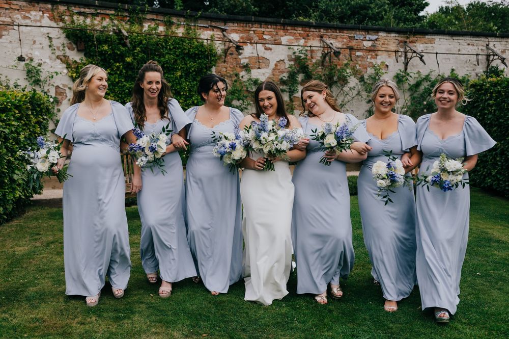 Wedding party in matching light blue dresses joyfully leaping in unison on a grassy lawn during outdoor celebration.