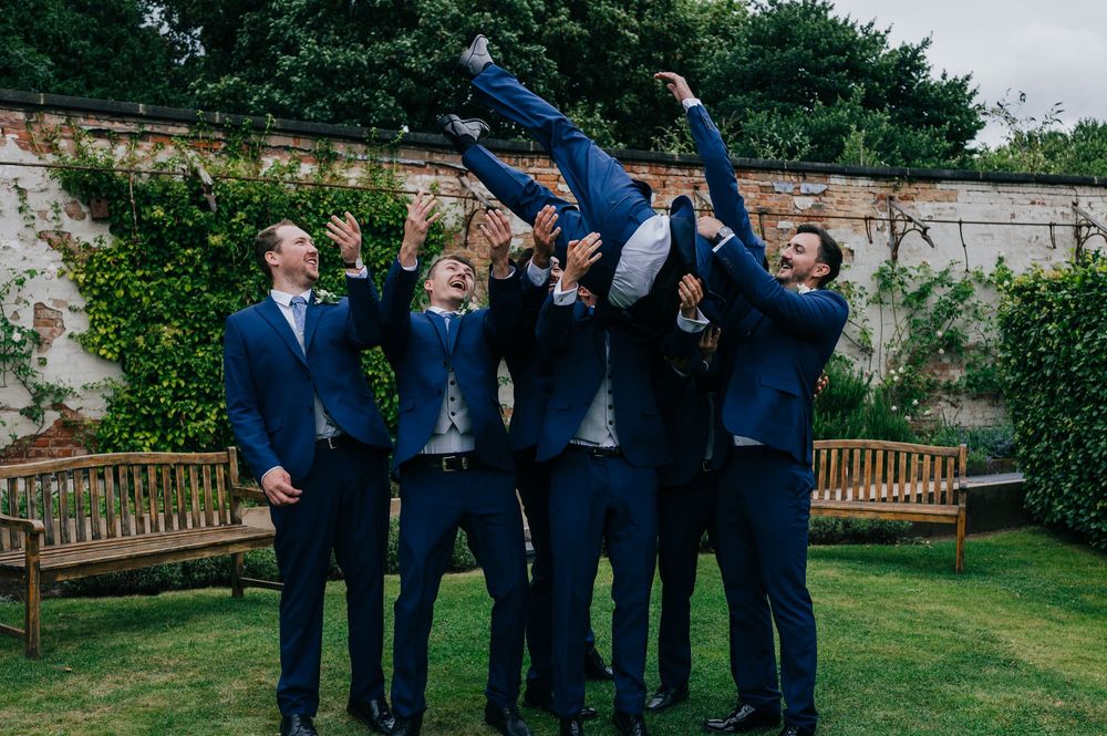 Groomsmen in navy blue suits playfully toss someone in the air during outdoor wedding photo session in a garden setting.