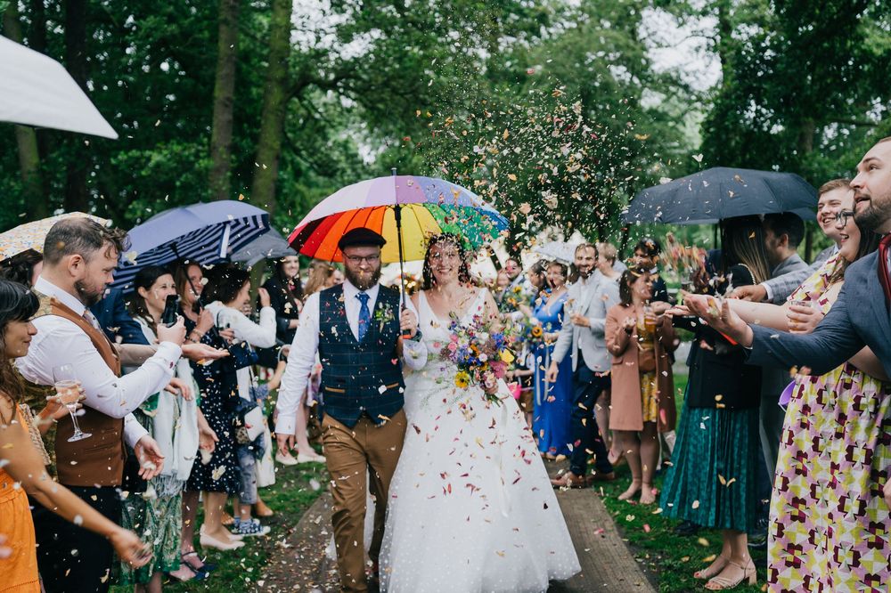 Wedding guests with colorful umbrellas shower confetti on newlyweds walking down pathway at outdoor forest ceremony.