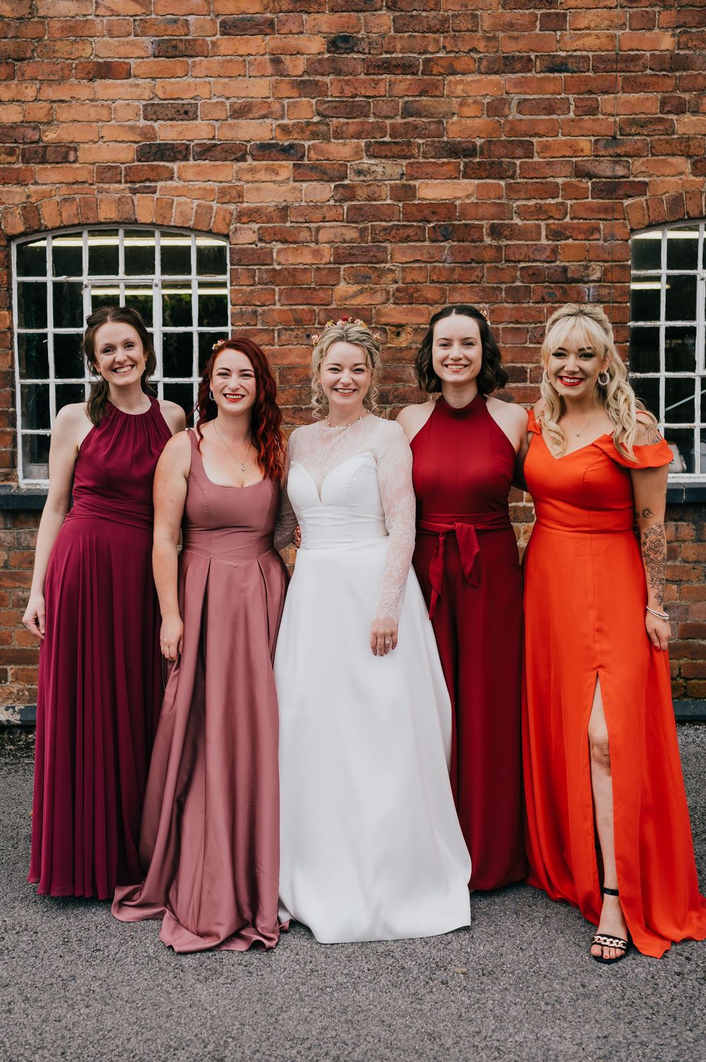 Bridesmaids in burgundy, dusty rose and bright orange dresses pose alongside bride in white gown against brick wall.