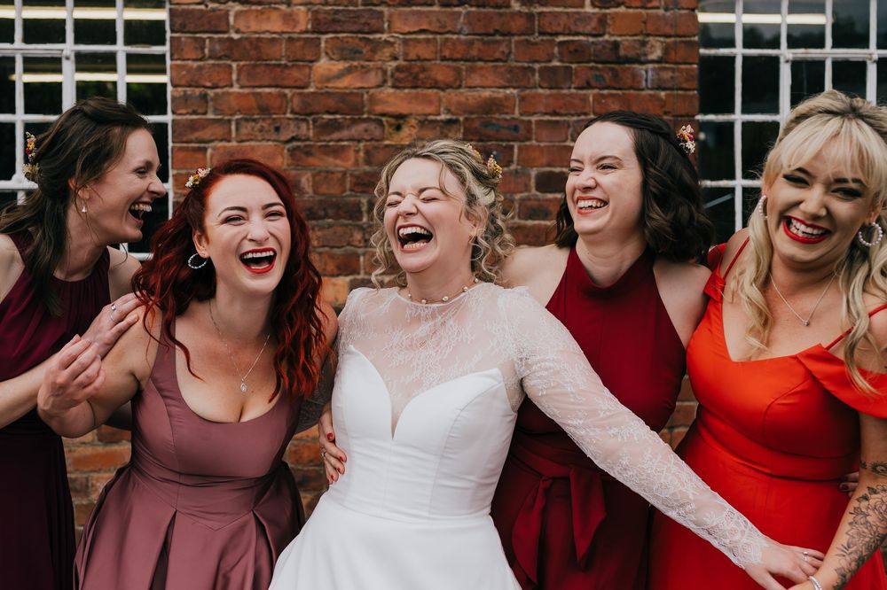 A joyful candid wedding party moment with five women sharing genuine laughter against a brick wall.