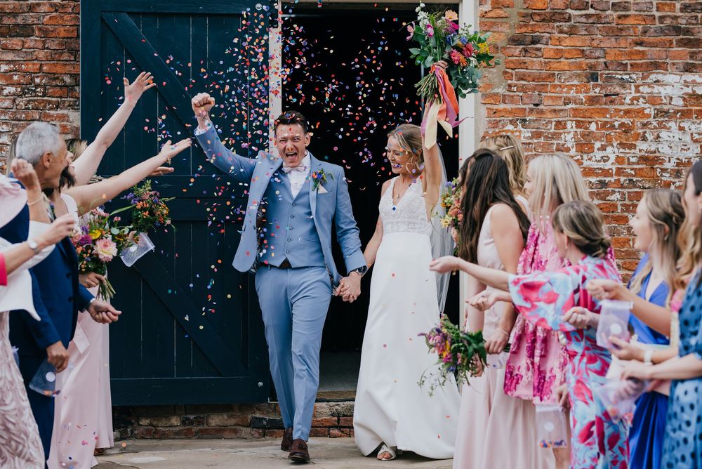 Wedding guests toss pink and white confetti as the newlyweds exit the rustic brick venue in celebration.