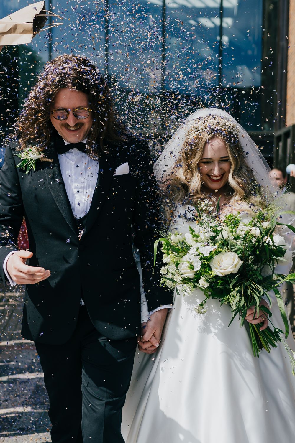 Newlyweds celebrate with confetti shower outside as bride holds white floral bouquet in traditional wedding attire.