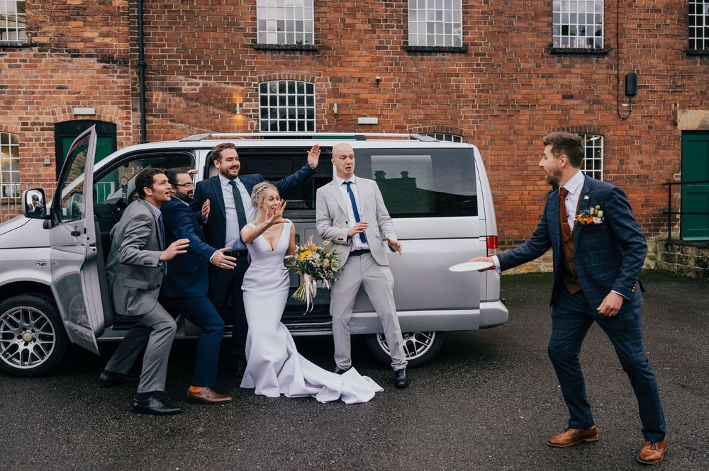 Wedding party poses informally by vintage vehicles outside brick industrial building while someone takes photos.
