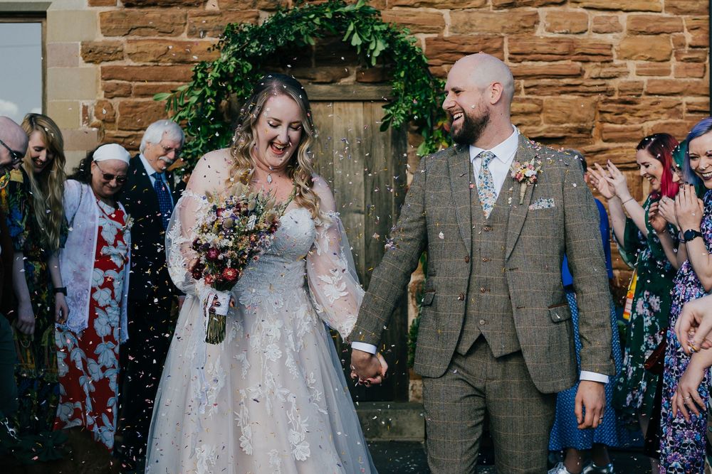 Wedding party celebrates with confetti outside rustic stone church venue during joyful celebration.