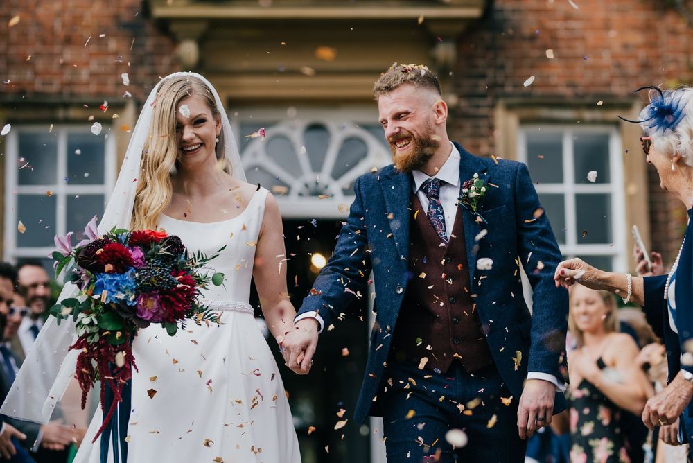The happy couple exits their wedding venue while guests shower them with confetti against a brick building backdrop.