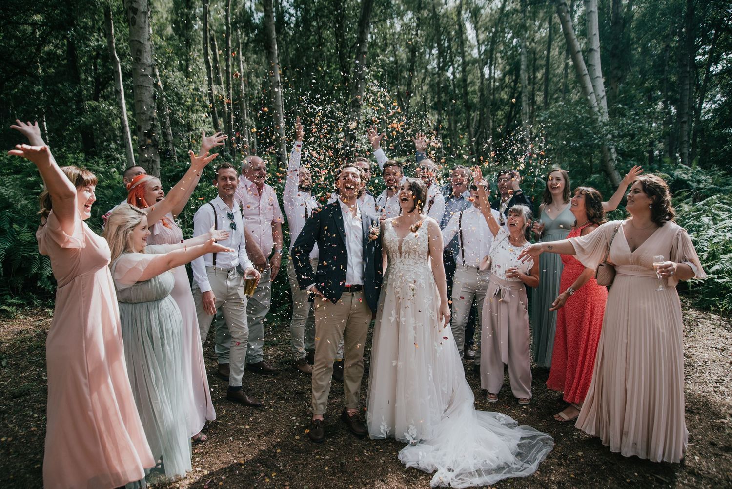 A joyful wedding party celebrates in a forest setting with bridesmaids in pastel dresses throwing confetti in the air.