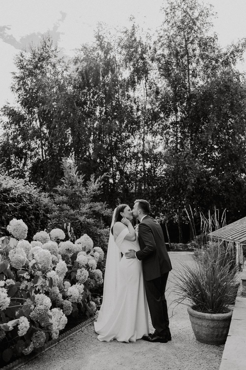 A couple shares a tender moment in a garden setting with lush trees and blooming hydrangeas in black and white.