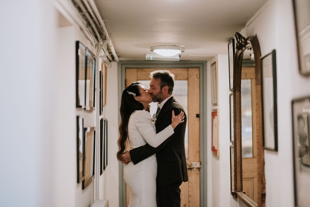 Romantic couple embraces and kisses in a narrow hallway with framed artwork on white walls.