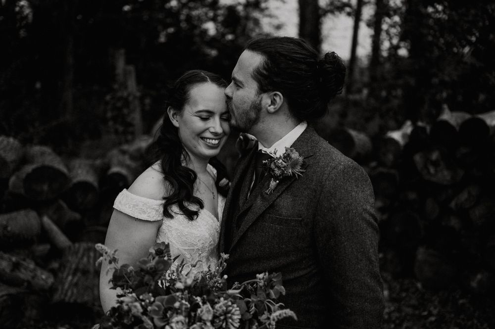 Black and white photo of a joyful couple sharing an intimate moment in the woods while holding a wedding bouquet.