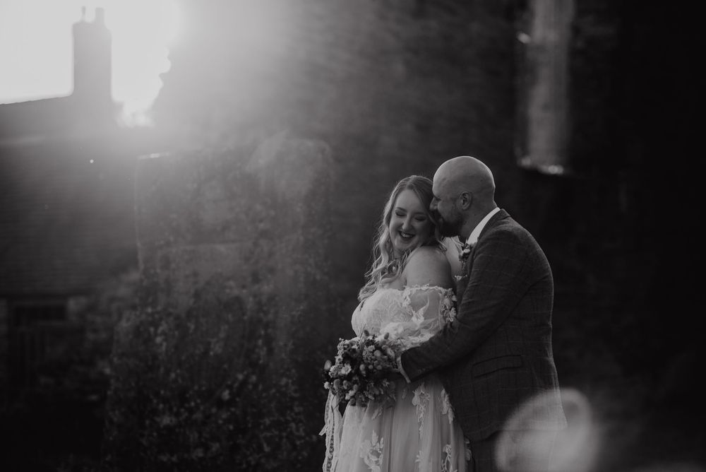 A romantic black and white moment of a bride and groom embracing in dramatic natural sunlight during their wedding ceremony.