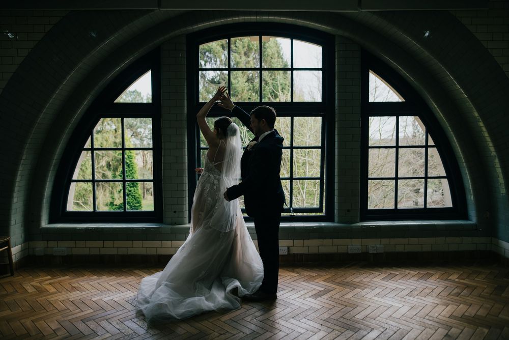 Two people dancing together in front of an arched window in a room with wooden flooring.