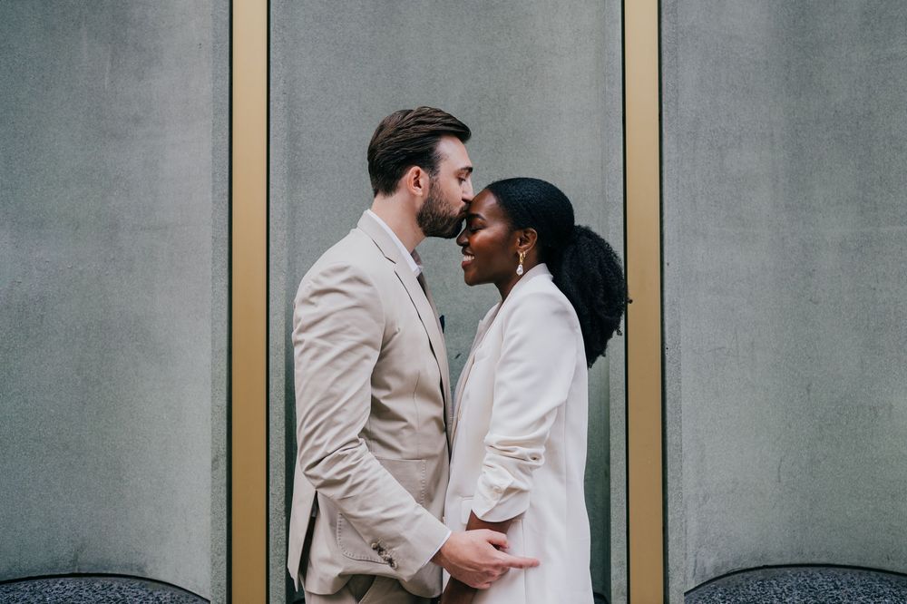 A romantic couple in matching light colored outfits share an intimate moment against a gray wall backdrop.