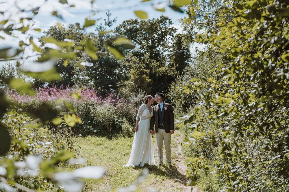 A wedding couple stands in a sunlit orchard surrounded by blooming trees on a beautiful summer day.