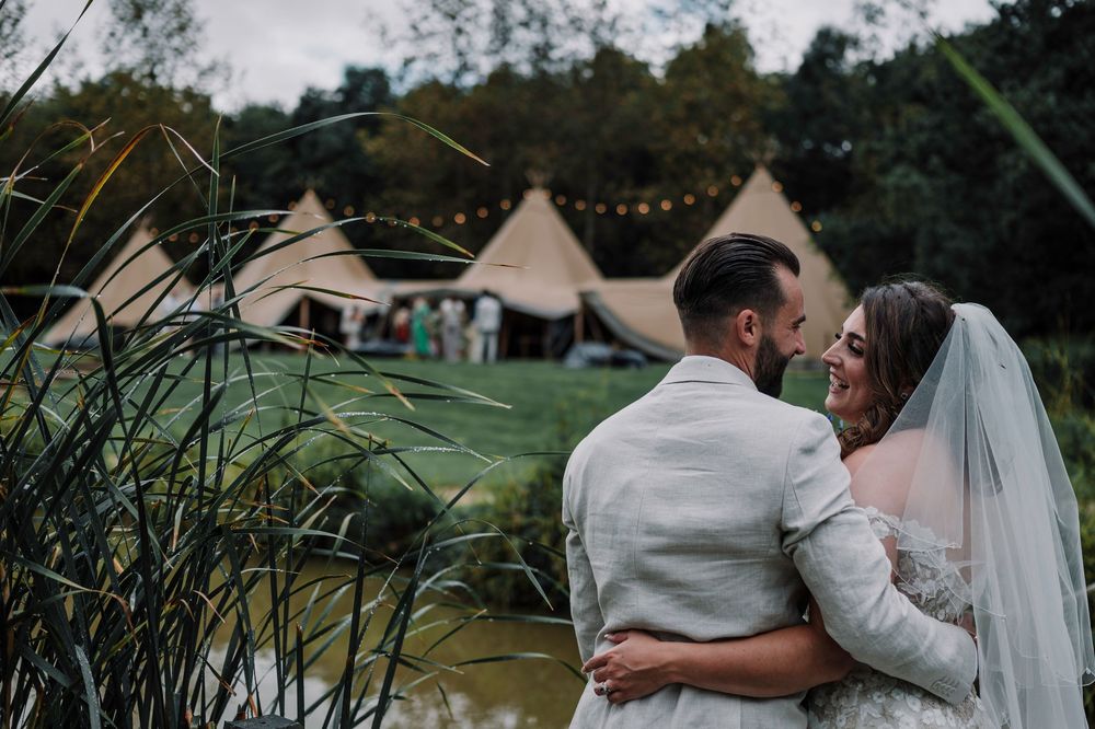 A couple in wedding attire embrace near a lake with tipi tents and string lights in the background at dusk.