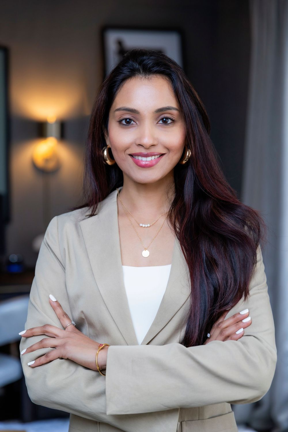 Professional headshot of someone in a beige blazer and white top standing with arms crossed in an elegant office setting.