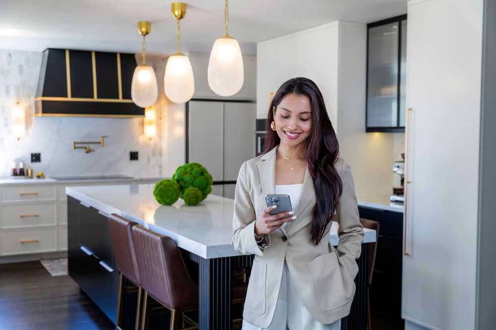 A person in a cream blazer smiles while holding a phone in an elegant modern kitchen with pendant lights.