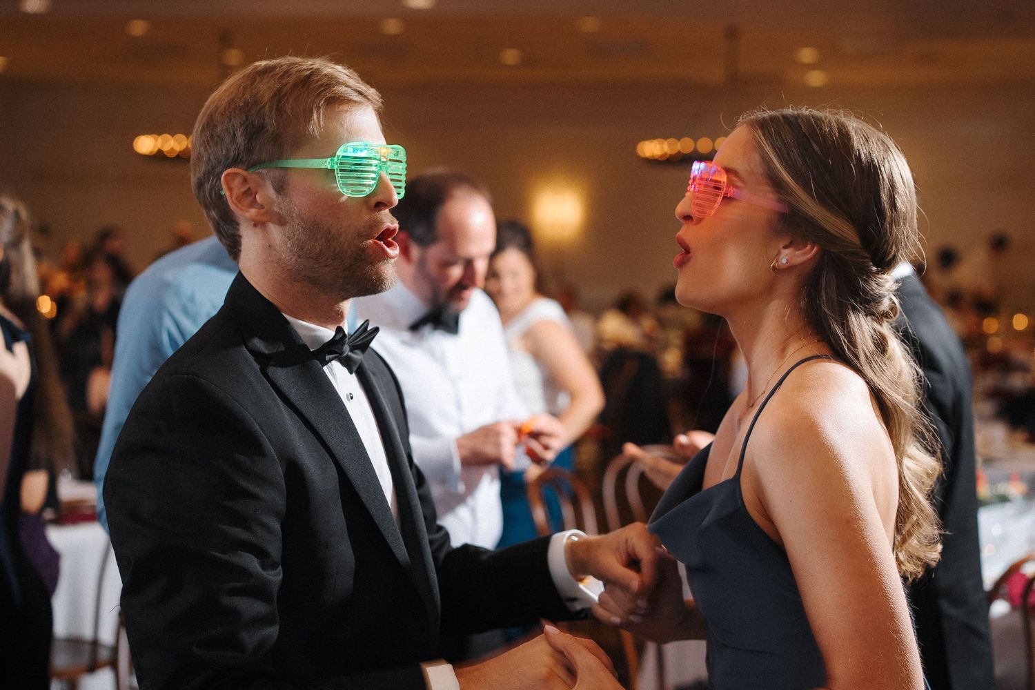 Two people wearing neon sunglasses dance and laugh together at a formal event with a reception hall in the background.
