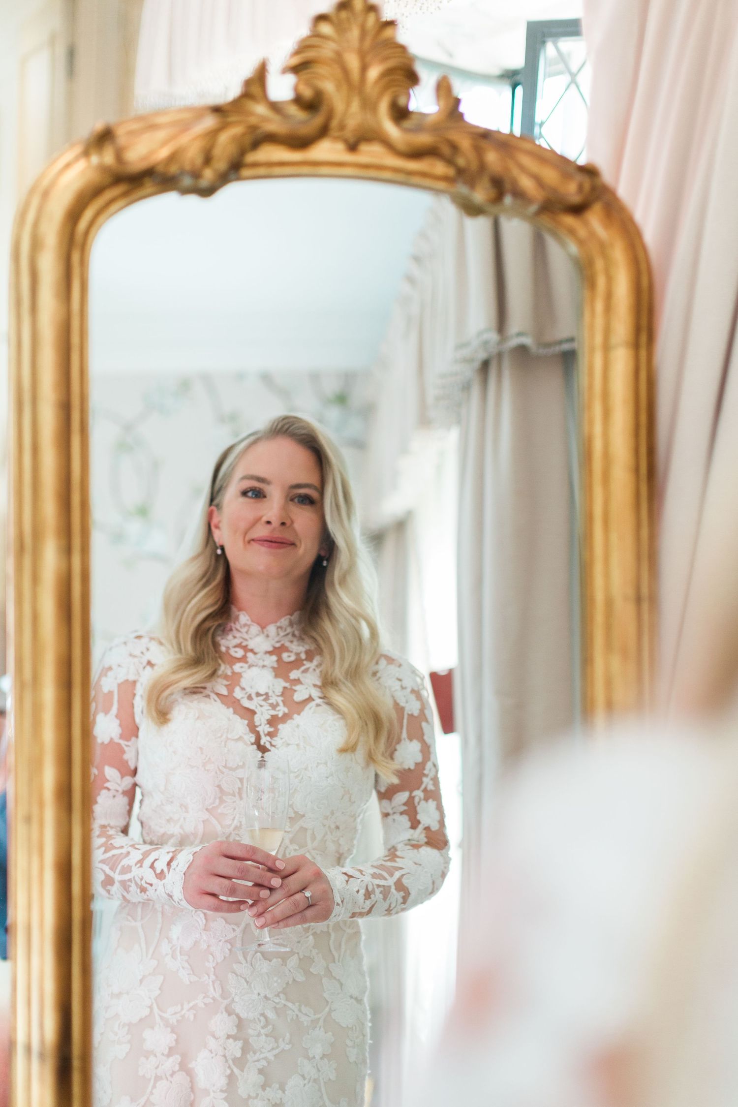 Bride wearing elegant lace wedding dress with long sleeves reflects in ornate gold vintage mirror with decorative frame at Ellenborough park