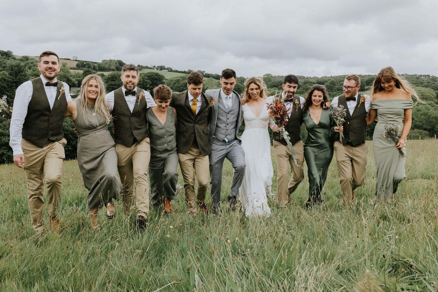 Wedding party in rustic attire poses together in a grassy field with rolling hills in the background.
