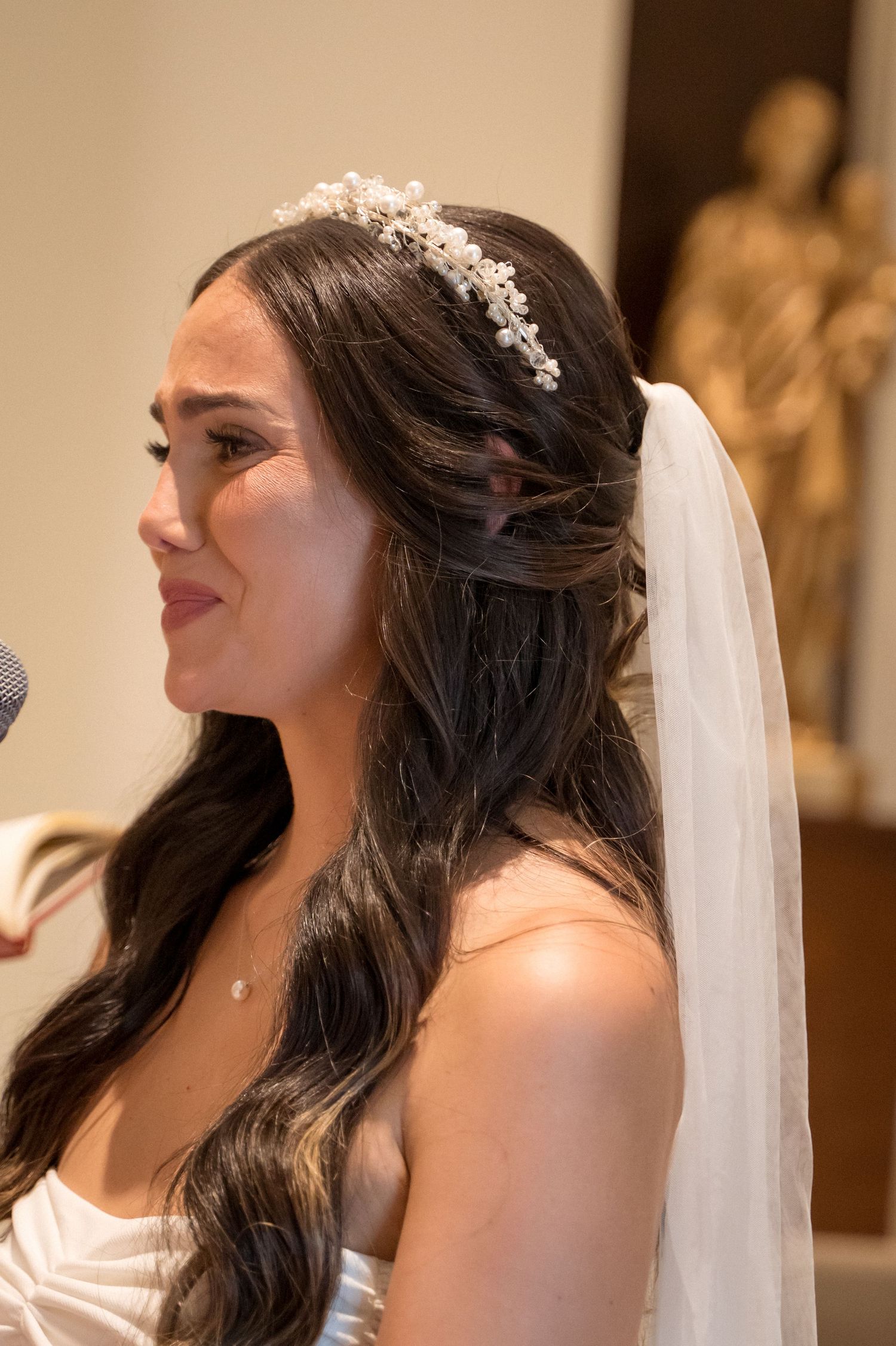 A bride wearing a delicate crystal headpiece and flowing veil smiles during a wedding ceremony.