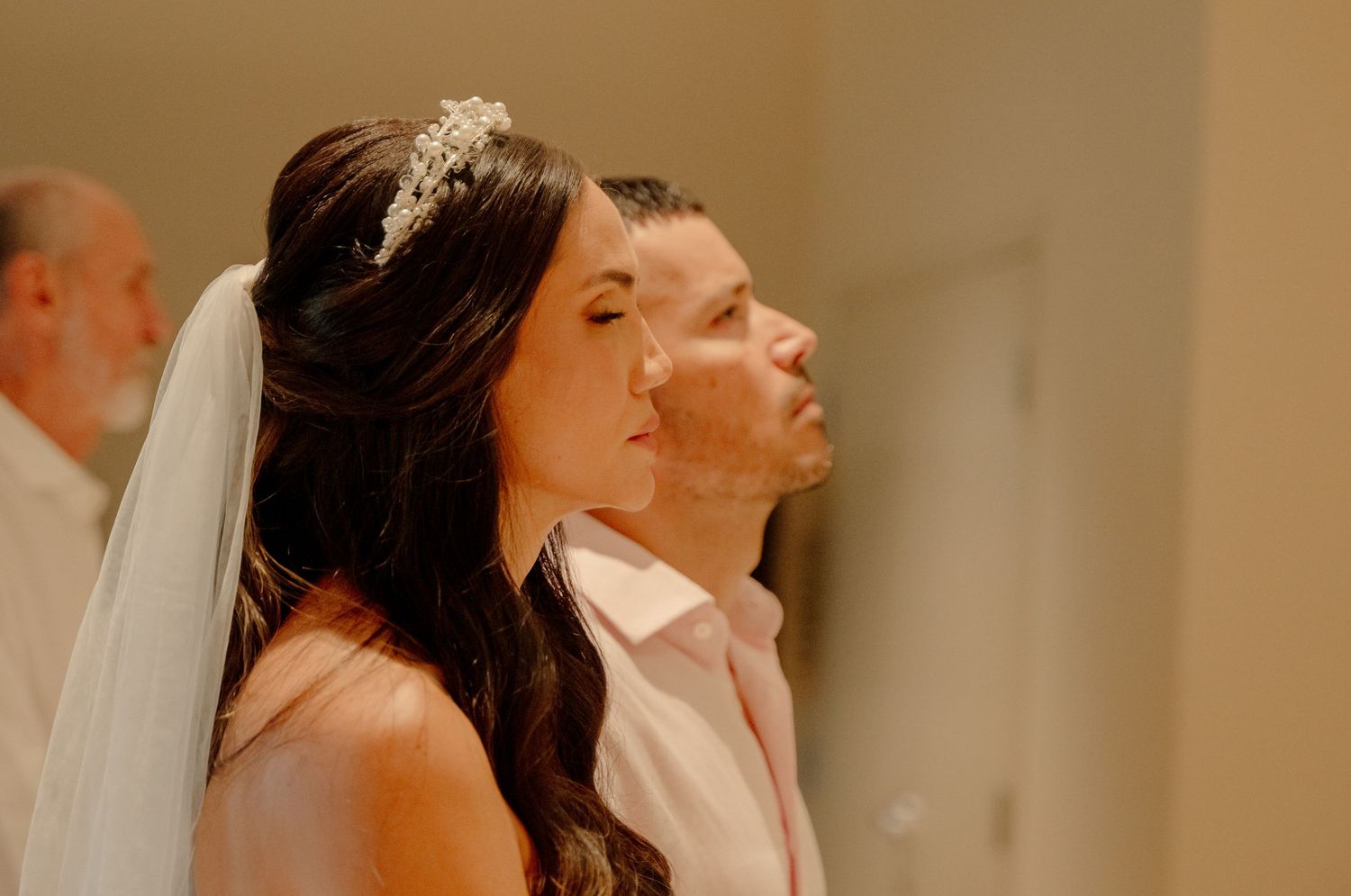 A couple in traditional wedding attire standing together during their ceremony in a warm-lit indoor setting.