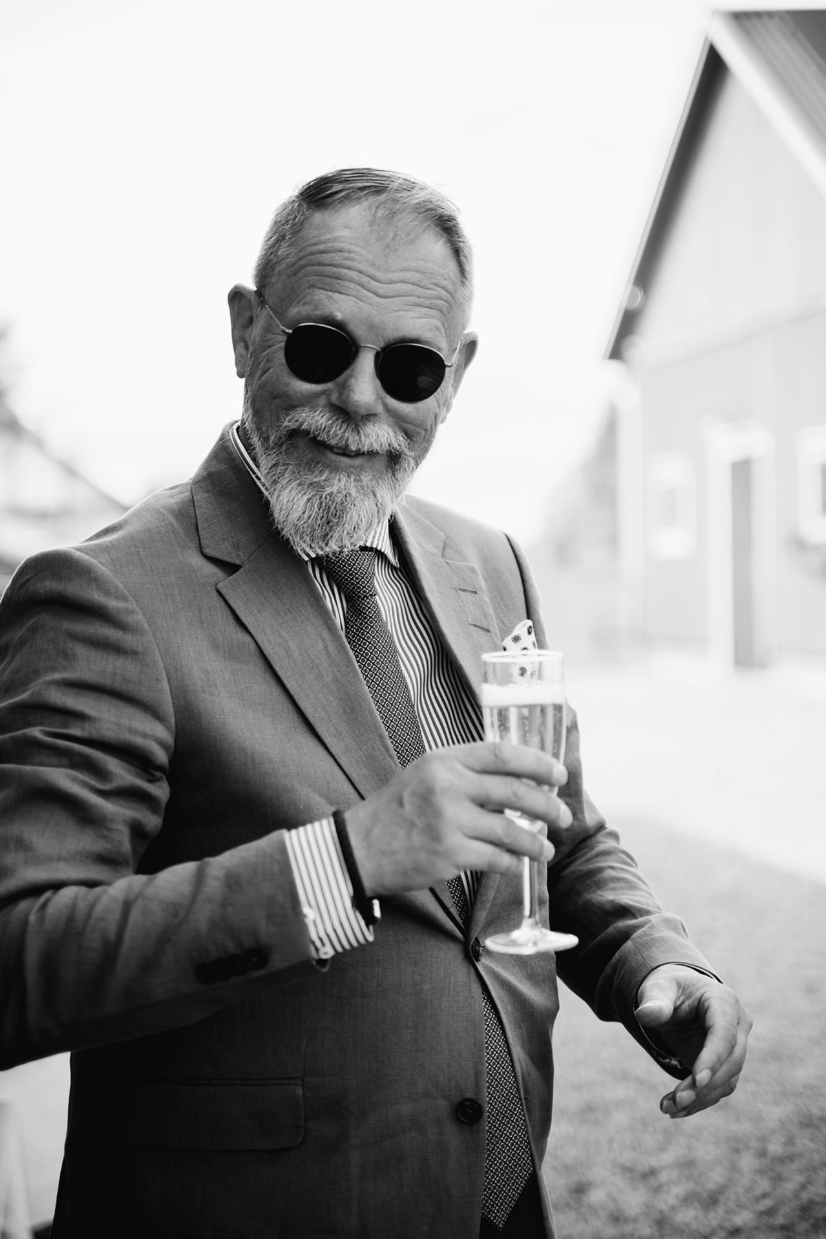 Bröllopsgäst i solgalsögon och ett glas bubbel i handen, mannen ler rakt in i kameran när han passerar förbi. Ett ögonblick fångad i farten.