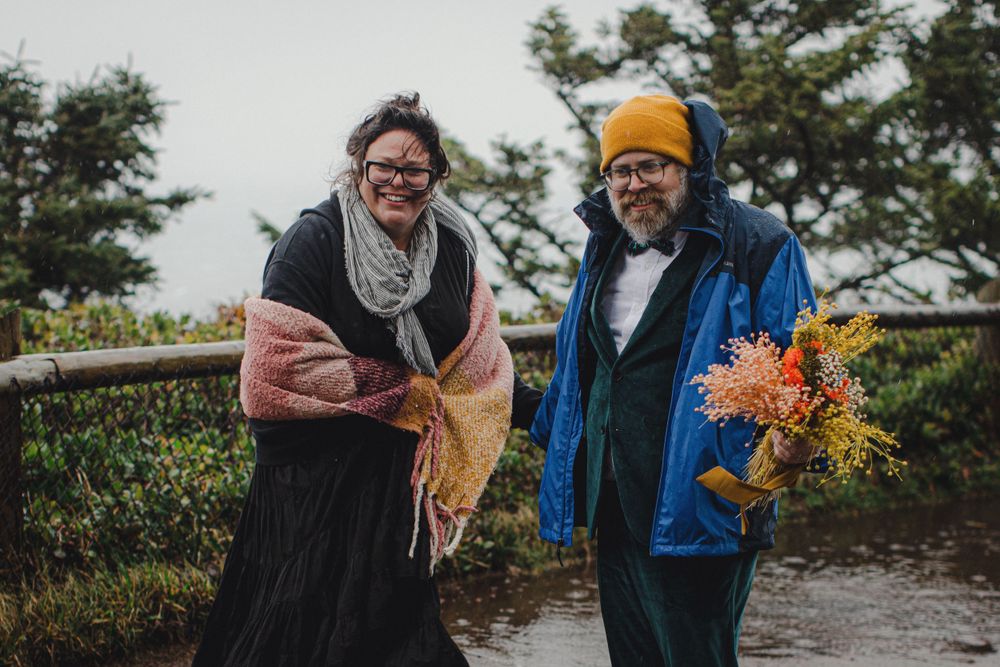 A pair in a blue coat and black dress share colorful flowers on a misty outdoor path.