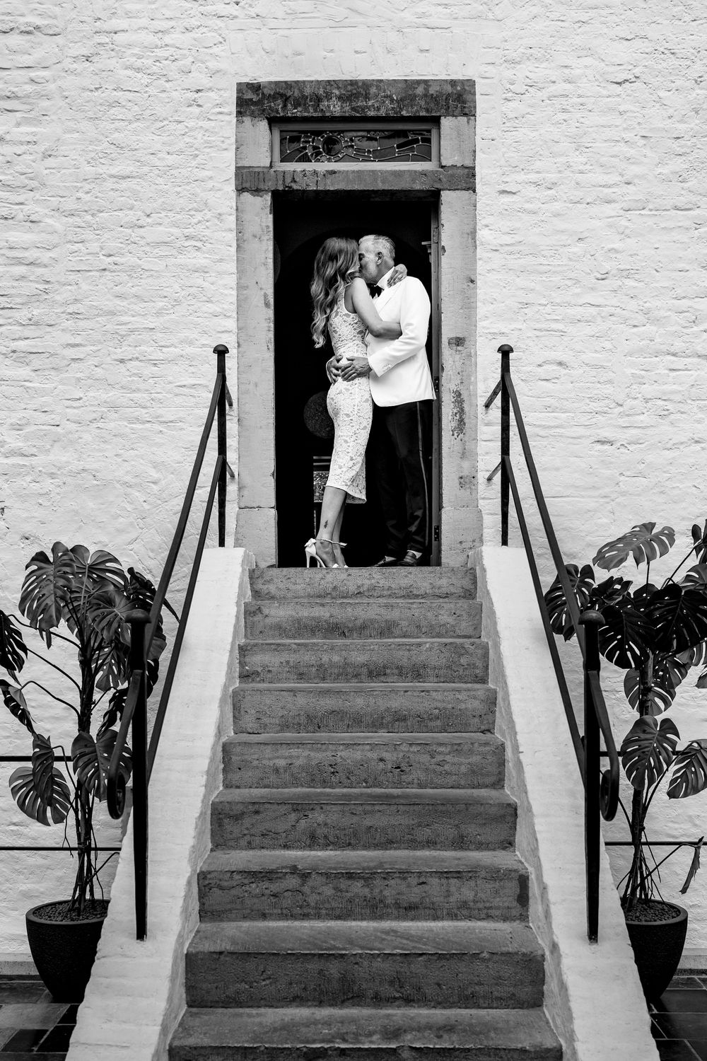 A romantic black and white photograph of a couple embracing in a doorway at the top of elegant stone steps with plants.