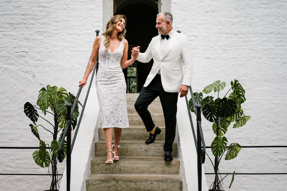 Newlyweds descend white stairs together with greenery decor at their wedding venue.