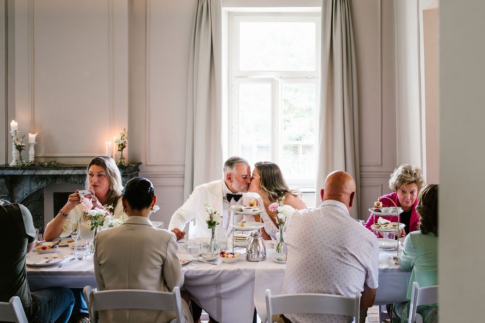 Wedding guests enjoy a formal dinner reception at an elegant table setting in a bright room.
