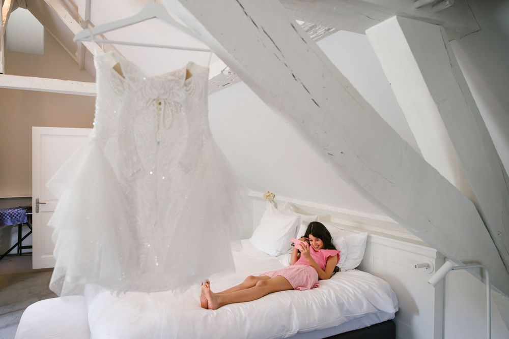 A white wedding dress hangs above a bed where someone rests during wedding preparations.
