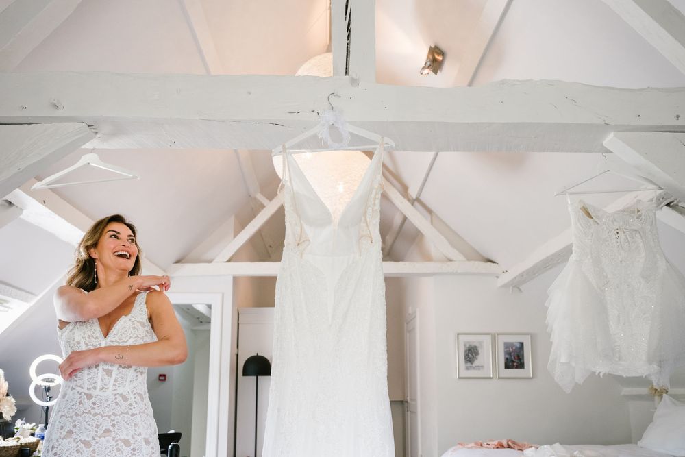Bride in white robe admires her wedding dress hanging in a bright white bridal suite.
