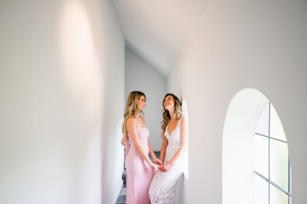 Two people in pink dresses share a tender moment by a bright window with an arched frame.