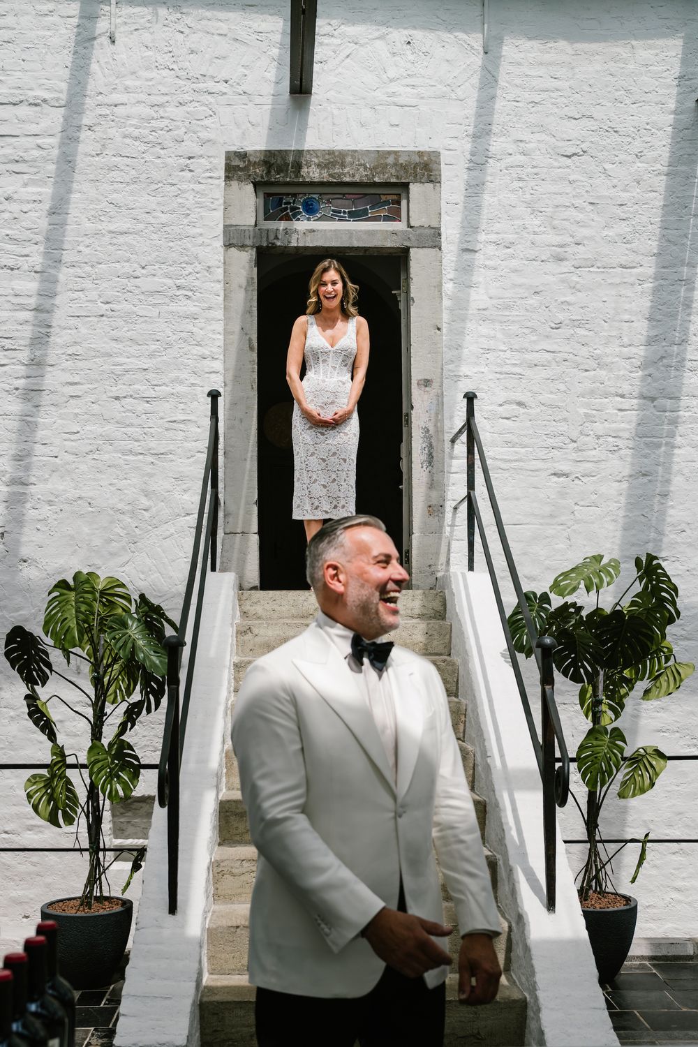 A first look photo of a groom and bride on white stairs surrounded by potted plants at an outdoor venue.